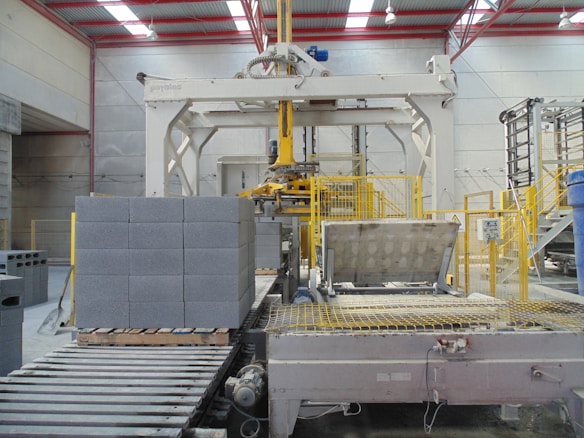 An industrial setting featuring a large machinery setup for handling cinder blocks. The foreground shows a stack of gray cinder blocks neatly arranged on a wooden pallet. Yellow safety railings and metal components are prominently visible. The area is well-lit by overhead lights and a structured metal framework forms the ceiling.