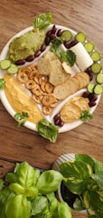 A platter of assorted foods including sliced cucumbers, soft cheese, pretzels, various types of spread such as avocado and hummus, olives, and crackers arranged on a white plate. Fresh herbs like basil and cilantro garnish the platter. The background is a wooden table, and in the foreground, there are potted basil and pothos plants.