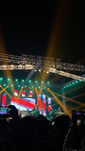 A vibrant music concert scene with a large stage illuminated by colorful lights. The background features an LED display with abstract red and blue graphics. Several musicians are performing, some playing guitars and drums. The audience, visible in silhouette, is engaged, taking photos and videos with their phones.