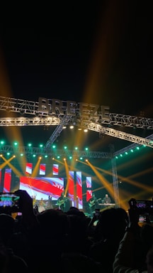 A vibrant music concert scene with a large stage illuminated by colorful lights. The background features an LED display with abstract red and blue graphics. Several musicians are performing, some playing guitars and drums. The audience, visible in silhouette, is engaged, taking photos and videos with their phones.
