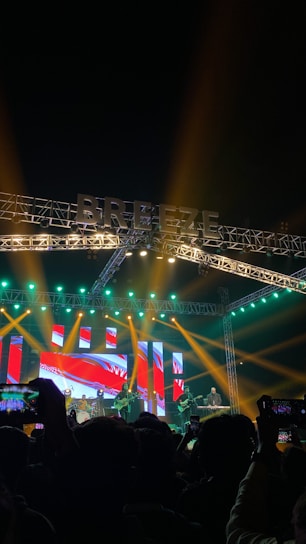 A vibrant music concert scene with a large stage illuminated by colorful lights. The background features an LED display with abstract red and blue graphics. Several musicians are performing, some playing guitars and drums. The audience, visible in silhouette, is engaged, taking photos and videos with their phones.