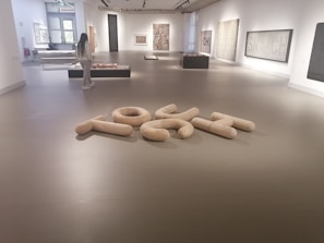A modern art gallery with various abstract artworks displayed on the walls and platforms. A person is observing the exhibits. The floor has a sculptural arrangement of large, light-colored, cushioned shapes forming an abstract composition. The space is well-lit with natural and artificial lighting.