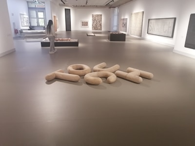 A modern art gallery with various abstract artworks displayed on the walls and platforms. A person is observing the exhibits. The floor has a sculptural arrangement of large, light-colored, cushioned shapes forming an abstract composition. The space is well-lit with natural and artificial lighting.