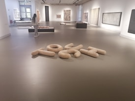 A modern art gallery with various abstract artworks displayed on the walls and platforms. A person is observing the exhibits. The floor has a sculptural arrangement of large, light-colored, cushioned shapes forming an abstract composition. The space is well-lit with natural and artificial lighting.