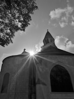 A black and white image showcasing a church with a tall, pointed steeple. Sunlight beams through, creating a starburst effect against the building's stone facade. A tree with dense foliage is visible on the left, and fluffy clouds fill the sky.