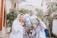 A group of young individuals dressed in white outfits, with two holding books titled 'Detik Detik' which partially cover their faces. They are standing outdoors, possibly in a garden or courtyard, with greenery and a few buildings in the background.
