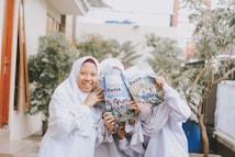 A group of young individuals dressed in white outfits, with two holding books titled 'Detik Detik' which partially cover their faces. They are standing outdoors, possibly in a garden or courtyard, with greenery and a few buildings in the background.