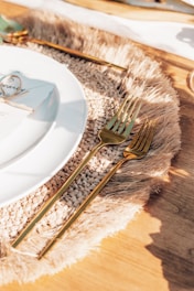 A close-up of a neatly arranged dining setting features a white plate with a menu card tied with a string on top. Accompanying the plate are gold-colored forks and knives placed on a decorative woven mat with fringed edges, all set on a wooden table surface.