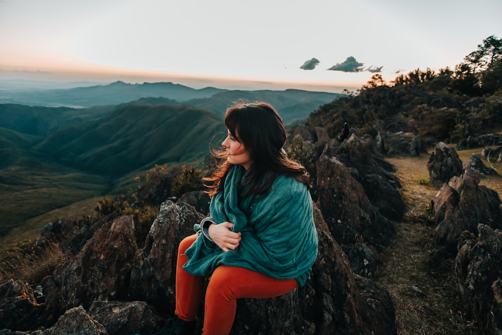 woman in teal long sleeve shirt and orange pants sitting on rock during daytime
