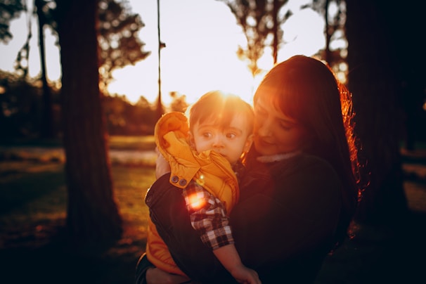 A mother holds her child close, enveloped in warm, golden sunlight filtering through surrounding trees. The child is dressed in a bright yellow jacket, looking curiously into the distance, while the mother gazes lovingly at the child. The background features soft focus silhouettes of trees under a sunset sky.