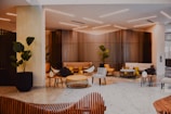 A modern indoor lounge area features stylish furniture including armchairs, stools, and couches in a mix of neutral and yellow hues. Wooden slatted walls create an elegant backdrop. Potted plants are strategically placed, adding greenery to the space, which is lit by sleek, minimalist ceiling lights. The floor is covered in large, light-colored tiles, while tables with minimalist designs add functionality to the seating area.
