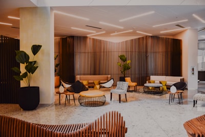 A modern indoor lounge area features stylish furniture including armchairs, stools, and couches in a mix of neutral and yellow hues. Wooden slatted walls create an elegant backdrop. Potted plants are strategically placed, adding greenery to the space, which is lit by sleek, minimalist ceiling lights. The floor is covered in large, light-colored tiles, while tables with minimalist designs add functionality to the seating area.
