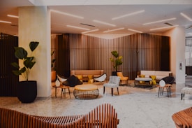 A modern indoor lounge area features stylish furniture including armchairs, stools, and couches in a mix of neutral and yellow hues. Wooden slatted walls create an elegant backdrop. Potted plants are strategically placed, adding greenery to the space, which is lit by sleek, minimalist ceiling lights. The floor is covered in large, light-colored tiles, while tables with minimalist designs add functionality to the seating area.