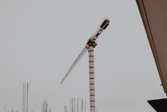 A construction crane is prominently displayed against a gray sky, with the framework of a building in the foreground. Steel reinforcement bars extend vertically, indicating ongoing construction work.