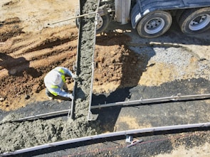 A construction worker is pouring concrete from a cement truck into a mold on a construction site. The area is surrounded by dirt and gravel, and the worker is wearing a high-vis vest and hard hat.