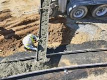 A construction worker is pouring concrete from a cement truck into a mold on a construction site. The area is surrounded by dirt and gravel, and the worker is wearing a high-vis vest and hard hat.