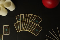 A fan of tarot-like cards is spread out on a dark surface, accompanied by three white candles on the left, a red sphere at the top right, and a few thin sticks at the bottom right.