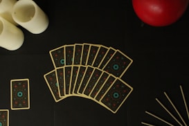 A fan of tarot-like cards is spread out on a dark surface, accompanied by three white candles on the left, a red sphere at the top right, and a few thin sticks at the bottom right.