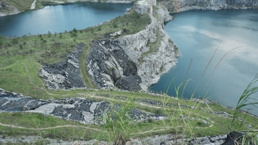A scenic landscape depicting a quarry with steep rocky cliffs and vibrant green grass surrounding a deep blue lake. The terrain appears rugged with exposed rock layers and some sparse vegetation growing on the slopes.