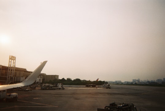 A view of an airport tarmac with the wingtip of an airplane visible in the foreground. Several airport vehicles and pieces of ground support equipment are scattered across the tarmac. Buildings are visible in the background, along with some trees. The sky is overcast, creating a subdued atmosphere.