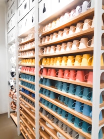 A neatly organized display of colorful ceramic mugs arranged on wooden shelves in a retail store setting. The mugs are set in rows, showcasing a spectrum of colors including browns, yellows, oranges, reds, blues, and whites. Above the shelves, images depicting different types of items suggest a sectioned layout or categorization. The lighting is bright, enhancing the vivid colors and creating a clean, modern look.