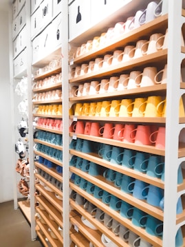 A neatly organized display of colorful ceramic mugs arranged on wooden shelves in a retail store setting. The mugs are set in rows, showcasing a spectrum of colors including browns, yellows, oranges, reds, blues, and whites. Above the shelves, images depicting different types of items suggest a sectioned layout or categorization. The lighting is bright, enhancing the vivid colors and creating a clean, modern look.