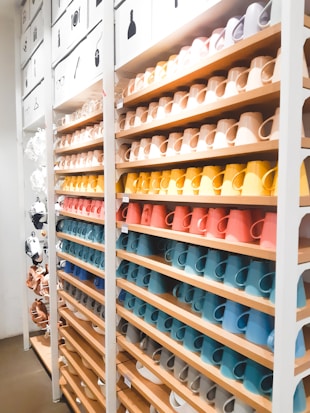A neatly organized display of colorful ceramic mugs arranged on wooden shelves in a retail store setting. The mugs are set in rows, showcasing a spectrum of colors including browns, yellows, oranges, reds, blues, and whites. Above the shelves, images depicting different types of items suggest a sectioned layout or categorization. The lighting is bright, enhancing the vivid colors and creating a clean, modern look.