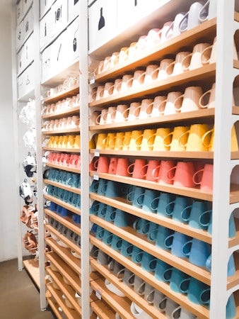 A neatly organized display of colorful ceramic mugs arranged on wooden shelves in a retail store setting. The mugs are set in rows, showcasing a spectrum of colors including browns, yellows, oranges, reds, blues, and whites. Above the shelves, images depicting different types of items suggest a sectioned layout or categorization. The lighting is bright, enhancing the vivid colors and creating a clean, modern look.