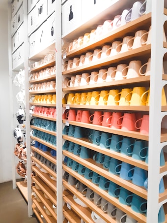 A neatly organized display of colorful ceramic mugs arranged on wooden shelves in a retail store setting. The mugs are set in rows, showcasing a spectrum of colors including browns, yellows, oranges, reds, blues, and whites. Above the shelves, images depicting different types of items suggest a sectioned layout or categorization. The lighting is bright, enhancing the vivid colors and creating a clean, modern look.