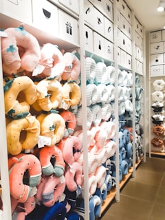 A store display features an assortment of colorful neck pillows arranged on shelves. The neck pillows come in various colors, including pink, yellow, blue, and white, and are shaped like animals or decorated with patterns. The background shows storage cabinets with simple black icons depicting different items. The shelves are neatly organized and well-lit, creating a bright and inviting shopping atmosphere.