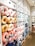 A store display features an assortment of colorful neck pillows arranged on shelves. The neck pillows come in various colors, including pink, yellow, blue, and white, and are shaped like animals or decorated with patterns. The background shows storage cabinets with simple black icons depicting different items. The shelves are neatly organized and well-lit, creating a bright and inviting shopping atmosphere.