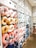 A store display features an assortment of colorful neck pillows arranged on shelves. The neck pillows come in various colors, including pink, yellow, blue, and white, and are shaped like animals or decorated with patterns. The background shows storage cabinets with simple black icons depicting different items. The shelves are neatly organized and well-lit, creating a bright and inviting shopping atmosphere.