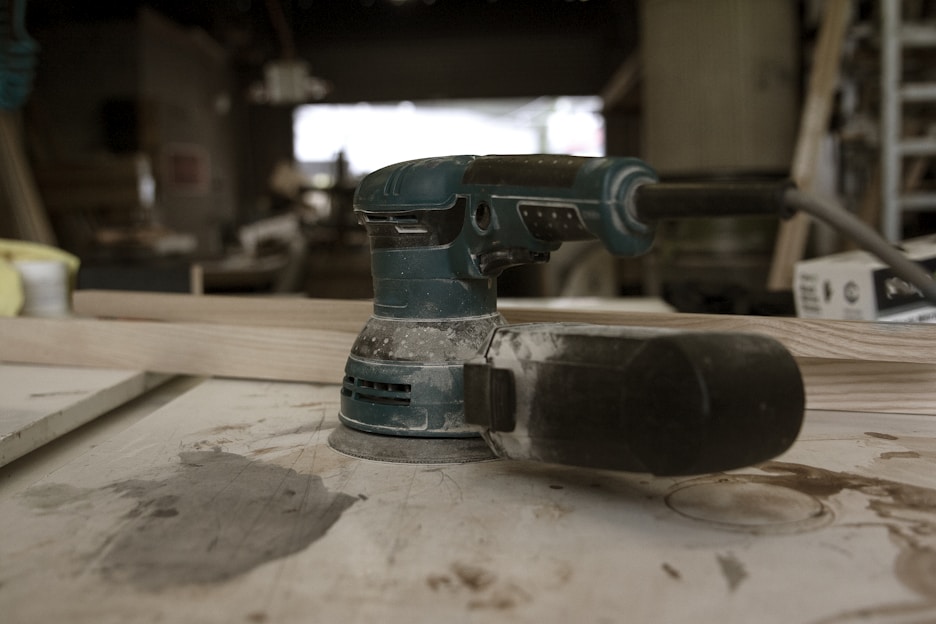 A power sander rests on a wooden workbench in a workshop, surrounded by wood shavings and other tools in the background. The sander appears to be in use, evident from the dust gathered on it. Ambient lighting suggests a calm and focused workspace.