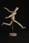 A wooden mannequin is posed dynamically on a dark background. It is supported by a metal rod, with its limbs arranged to suggest movement or a running action. The lighting highlights the natural wood tone of the figure, creating a sense of motion against the contrasting backdrop.
