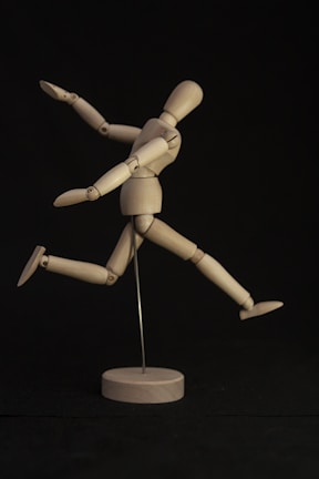 A wooden mannequin is posed dynamically on a dark background. It is supported by a metal rod, with its limbs arranged to suggest movement or a running action. The lighting highlights the natural wood tone of the figure, creating a sense of motion against the contrasting backdrop.