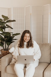 A person with long hair wearing a white outfit is sitting on a beige sofa using a laptop. The setting includes a potted plant to the left and a folding screen in the background, suggesting a cozy and private indoor space.