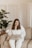 A person with long hair wearing a white outfit is sitting on a beige sofa using a laptop. The setting includes a potted plant to the left and a folding screen in the background, suggesting a cozy and private indoor space.
