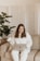 A person with long hair wearing a white outfit is sitting on a beige sofa using a laptop. The setting includes a potted plant to the left and a folding screen in the background, suggesting a cozy and private indoor space.