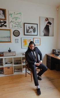 A well-dressed man in a suit sits confidently in a chair within a stylish, art-filled room. The walls are adorned with various framed photographs and artwork. In the background, shelves hold books and a vintage record player stands to the side.