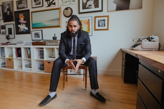 A man in a tailored suit sits confidently on a chair in a stylish, modern room. The background features a wall adorned with various framed artworks and photographs. On one side, a shelving unit contains books and baskets, while a vintage typewriter sits on a wooden table to the right.