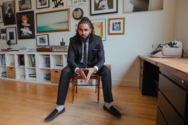 A man in a tailored suit sits confidently on a chair in a stylish, modern room. The background features a wall adorned with various framed artworks and photographs. On one side, a shelving unit contains books and baskets, while a vintage typewriter sits on a wooden table to the right.