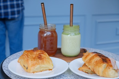 Two jars with metal straws contain beverages, one being a brown iced tea or coffee and the other a light green smoothie. They are placed on a wooden board. In front, two pastries rest on separate white plates, a pain au chocolat and a croissant. The background is light blue and blurred, and a person in a plaid shirt and jeans is partially visible.
