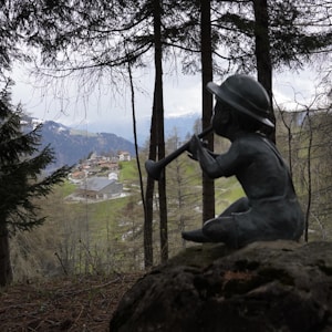 A serene pastoral landscape with a sculpture of a child sitting on a rock, playing a wind instrument. The foreground features tall trees, partially obscuring a distant view of a quaint mountain village nestled in a lush, green valley with snow-capped mountains in the background.