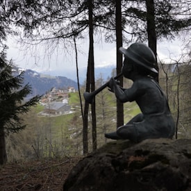 A serene pastoral landscape with a sculpture of a child sitting on a rock, playing a wind instrument. The foreground features tall trees, partially obscuring a distant view of a quaint mountain village nestled in a lush, green valley with snow-capped mountains in the background.