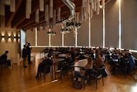 Attendees are seated in a spacious conference room with a high wooden ceiling and tubular light fixtures. A speaker at the front is presenting next to a projector screen, and the audience is attentively watching. Decorative elements hang from the ceiling, adding a festive touch.