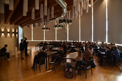 Attendees are seated in a spacious conference room with a high wooden ceiling and tubular light fixtures. A speaker at the front is presenting next to a projector screen, and the audience is attentively watching. Decorative elements hang from the ceiling, adding a festive touch.