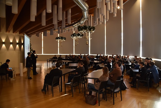 Attendees are seated in a spacious conference room with a high wooden ceiling and tubular light fixtures. A speaker at the front is presenting next to a projector screen, and the audience is attentively watching. Decorative elements hang from the ceiling, adding a festive touch.