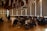Attendees are seated in a spacious conference room with a high wooden ceiling and tubular light fixtures. A speaker at the front is presenting next to a projector screen, and the audience is attentively watching. Decorative elements hang from the ceiling, adding a festive touch.