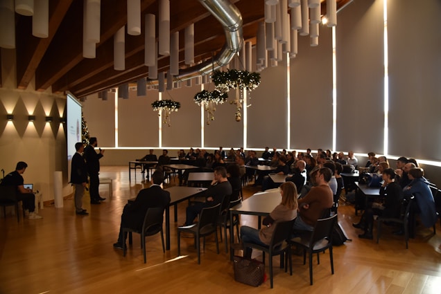 Attendees are seated in a spacious conference room with a high wooden ceiling and tubular light fixtures. A speaker at the front is presenting next to a projector screen, and the audience is attentively watching. Decorative elements hang from the ceiling, adding a festive touch.