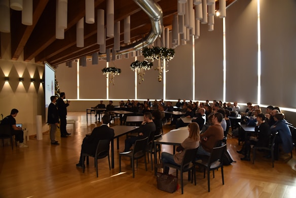 Attendees are seated in a spacious conference room with a high wooden ceiling and tubular light fixtures. A speaker at the front is presenting next to a projector screen, and the audience is attentively watching. Decorative elements hang from the ceiling, adding a festive touch.